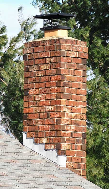 Nice red brick and mortar chimney with shingled roof.  In the background there a spruce trees and a light blue sky.