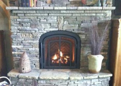 Mendota Gas Fireplace with arched insert. The suround hearth and mantel are stone. The mantel is decorated with books and leaves and vases.